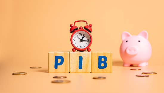 The word PIB in Brazilian Portuguese which is the abbreviation of the word gross domestic product, written on wooden cubes. A red alarm clock and coins from Brazil in the composition.