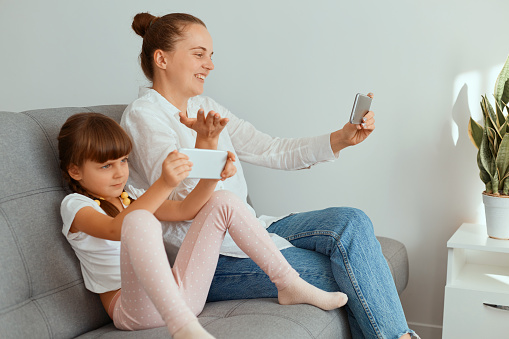 Side view portrait of smiling woman woman with hair bun wearing white shirt and jeans sitting with her cute kid on cough and having video call, expressing hand aside.