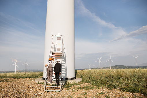 African american inspector in black suit and white helmet talking with indian technician in uniform while doing examining of windmill farm. Concept of people, inspection and clean energy.