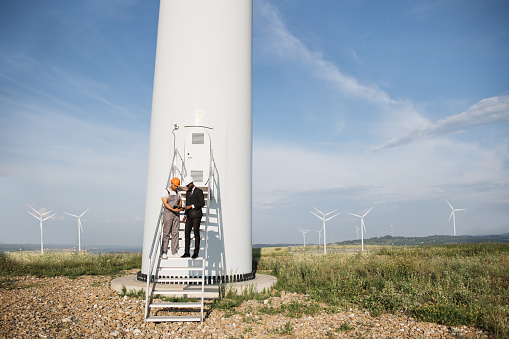 Multiracial partners standing on stairs near wind turbine and looking on screen of digital tablet. Two industrial workers turning on system operation of eco farm using modern gadgets.