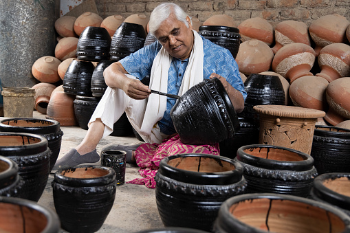 Happy Indian senior man of potters painting clay pots at workshop