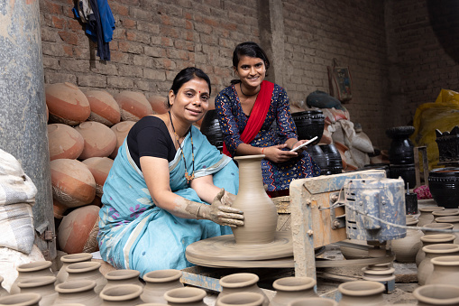 Mother and daughter using digital tablet while making clay pot on spinning wheel at village