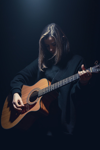 A young woman with an acoustic guitar on a stage in the dark under a ray of light, concert,, copy space.