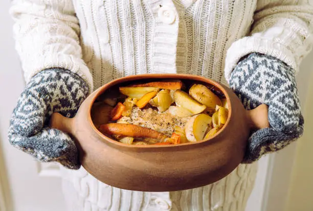 Woman holding terracotta clay cooking pot with slow cooked pork roast and vegetables inside. Wearing knitted clothing, winter comfort food concept.