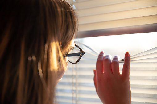 A woman with glasses looks through the blinds at the early morning sunlight.