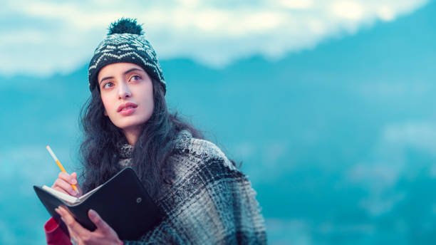 In this close-up, outdoor image with copy space, an Asian/Indian, beautiful young woman writes in a diary standing on the roof of her house against the beautiful scenic view of the mountains of Himachal Pradesh. She wears a warm cap and shawl.