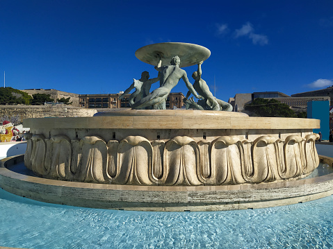 The Tritons’ Fountain (Maltese: Il-Funtana tat-Tritoni) is a fountain located just outside the City Gate of Valletta, Malta. It consists of three bronze Tritons holding up a large basin, balanced on a concentric base built out of concrete and clad in travertine slabs. The fountain is one of Malta's most important Modernist landmarks