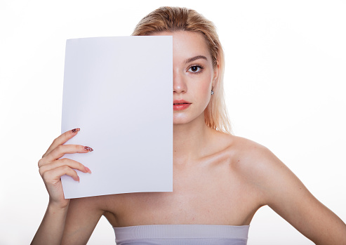 From a blank sheet of paper concept. Young woman holding white paper covering half of face looking at camera standing over white studio background. Female beauty portrait with copy space.
