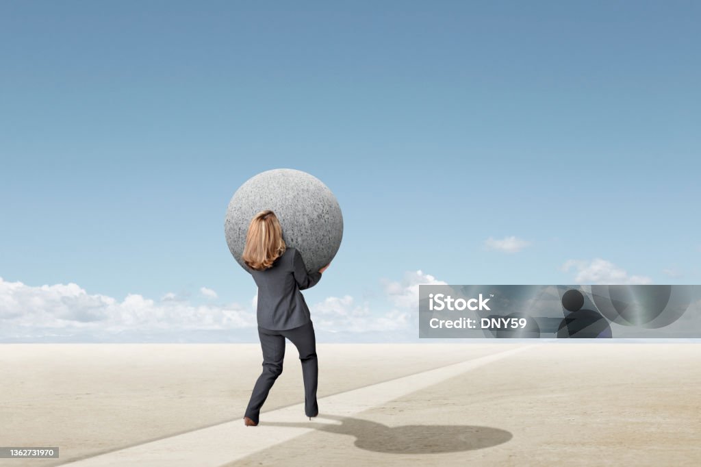 Woman Struggling To Carry Large Round Sphere Toward The Horizon A businesswoman leans back as she struggle to carry a large round sphere on her trek toward the horizon. Over-Burdened Stock Photo