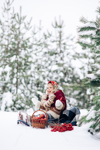 Beautiful little child girl sits with bagels bunch on her neck near basket of apples and viburnum bunches during the walk in snowy forest. She dressed in the old Russian style in red headscarf.