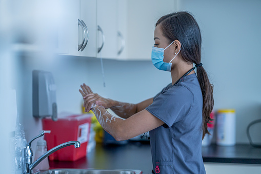 A nurse wearing a face mask washes her hands thoroughly in the sink of the doctor's office.