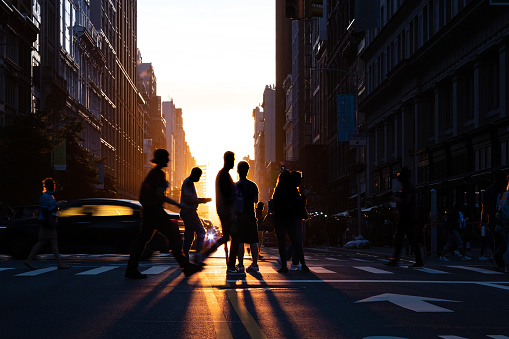 Silhouettes of people crossing a busy intersection on 5th Avenue in New York City with the light of sunset in the background