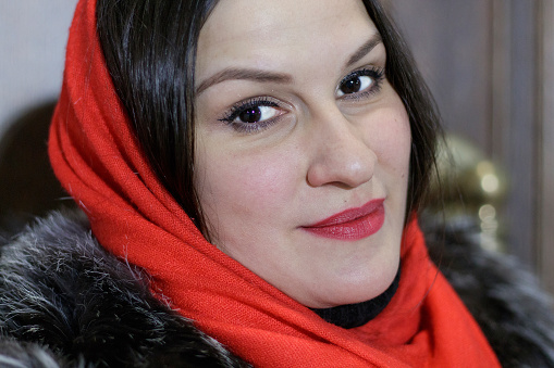 Close-up portrait of attractive Asian young woman with dark hair and brown eyes, wearing a red headscarf and fur coat, with red lipstick and glitter on her eyes, looking at the camera.