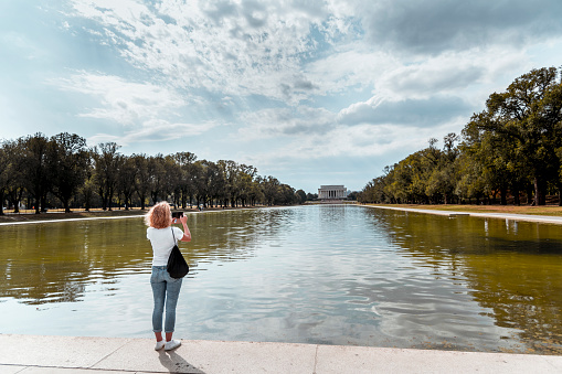 A tourist standing in front of the Reflecting Poll photographs the Lincoln Memorial on the end of it with her phone.