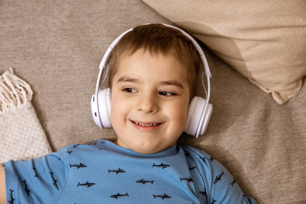 piccolo ragazzo caucasico con camicia blu e cuffie bianche che ascolta musica o audiolibro sul divano di casa. bambino carino che si rilassa, riposa nella sua stanza. - one little boy audio foto e immagini stock