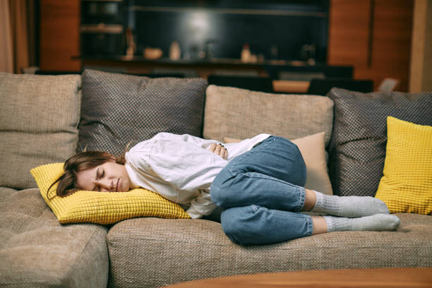 giovane donna malsana che soffre di dolore mestruale, tenendo la pancia, avendo crampi addominali durante il periodo sdraiato sul divano - pms foto e immagini stock