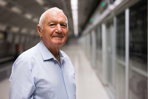 Portrait of positive European old man standing in subway station and waiting for train.