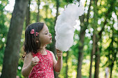 Little cute girl 3-4 eating cotton candy in sunny park, among tall trees on green grass
