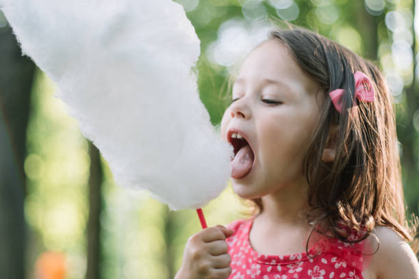 Little cute girl 3-4 eating cotton candy in sunny park, among tall trees on green grass stock photo