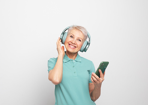 Happy mature short haired woman smiling broadly posing against whte grey background in wireless headphones, jolding modern smartphone, enjoying clean high quality sound, listening to music online