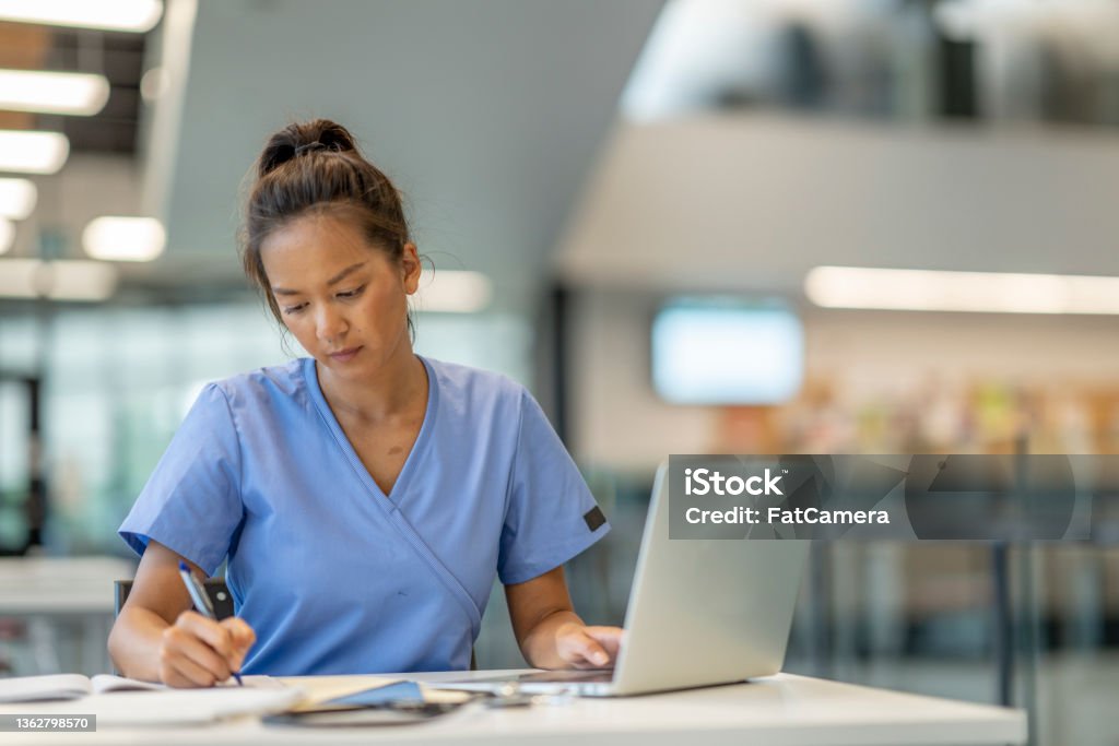 Medical Student Studying A female medical student sits at a table with her laptop out in front of her as she studies for class.  She is wearing blue scrubs and focused on the task. Medical Student Stock Photo
