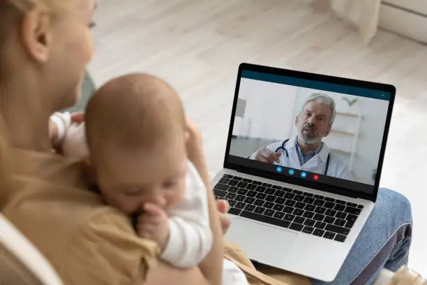 Photo of Mother holding baby, using laptop, making video call to pediatrician