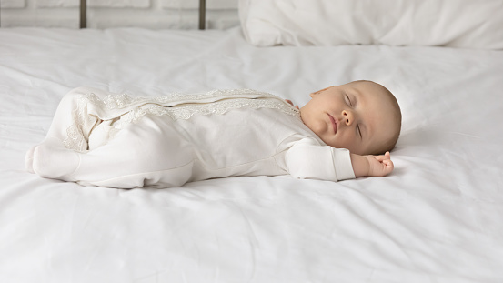 Cute baby in white bodysuit sleeping, adorable newborn child infant with closed eyes lying on back on comfortable bed white sheet, relaxing resting alone, fall asleep, childhood concept