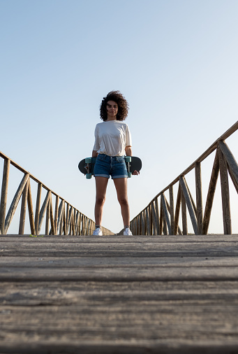 Young skater woman with afro hair standing holding her skateboard. Low angle view.