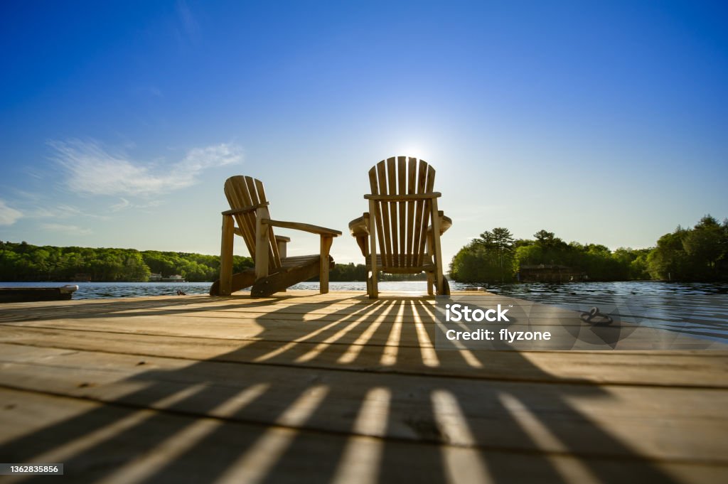 Sunrise on two empty Adirondack chairs sitting on a dock Cottage life - Sunrise on two empty Adirondack chairs sitting on a dock on a lake in Muskoka, Ontario Canada. The sun rays create long shadows on the wooden pier. Retirement Stock Photo