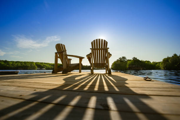 Sunrise on two empty Adirondack chairs sitting on a dock Cottage life - Sunrise on two empty Adirondack chairs sitting on a dock on a lake in Muskoka, Ontario Canada. The sun rays create long shadows on the wooden pier. retirement stock pictures, royalty-free photos & images