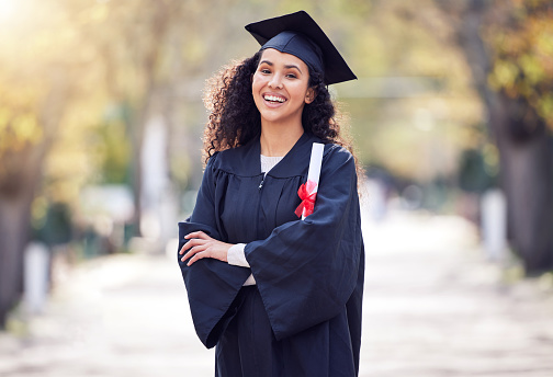 A smiling female graduate  happily holding her graduation certificate in her hands as she stands on the university steps wearing her gown and mortarboard, in the background her classmates are also holding their diplomas