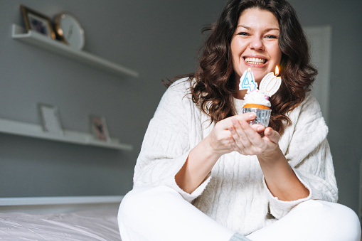 Young adult happy beautiful woman forty years plus size body positive in white cozy knitted sweater with festive cupcake with candle in hands on bed at home party, birthday girl