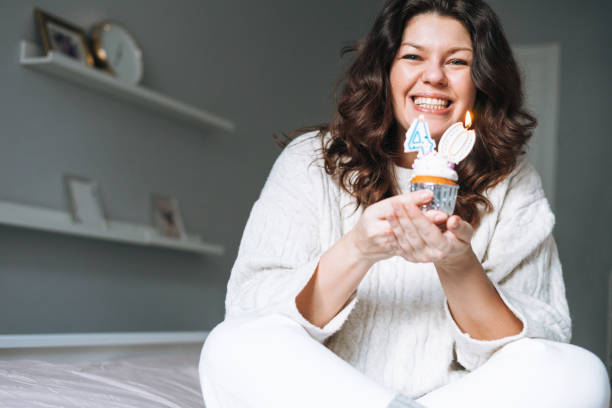 young adult happy beautiful woman forty years plus size body positive in white cozy knitted sweater with festive cupcake with candle in hands on bed at the home party, birthday girl - 40 44 years imagens e fotografias de stock