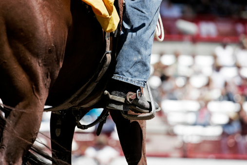 A cowboy waits on his horse inside a large rodeo arena during a daytime performance, with the grandstands in the background (shallow focus).