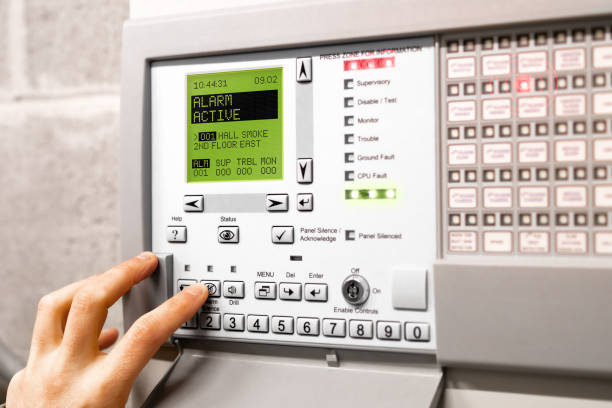 Fire alarm control panel is activated and in alert mode. Display message: Alarm active hall smoke. Red flickering lights and peeping. A hand is using the silence button. Selective focus. fire alarm photos stock pictures, royalty-free photos & images