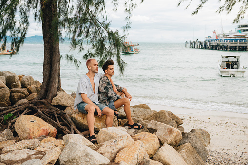 Gay Couple spend time together outdoors. Homosexual couple sitting on the stoned at the beach
