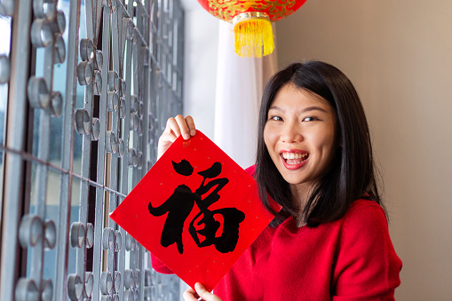 Asian woman celebrating chinese new year, wearing red dress holding couplet 'prosperity' (Chinese blessing word) at home