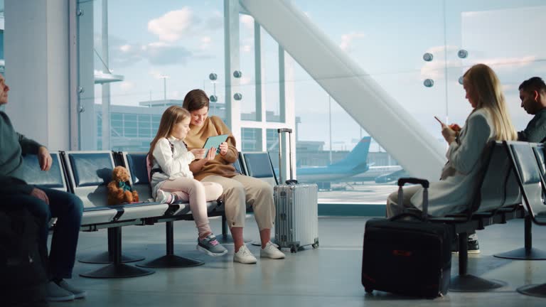 Busy Airport Terminal: Cute Mother and Little Daughter Wait for their Vacation Flight, Play Educational Games on Digital Tablet. People Sitting in Boarding Lounge of Airline Hub with Airplanes Flying