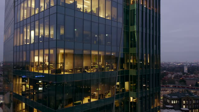 Drone shot at a night skyscraper with offices and people working late solving business issues and sitting at computers. Drone flies around the glass skyscraper from right to left.