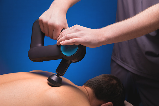 Close-up of a professional male masseur stimulates the back muscles of a male patient in a dark spa room for massage. Percussion mechanical effect on overstrained muscles