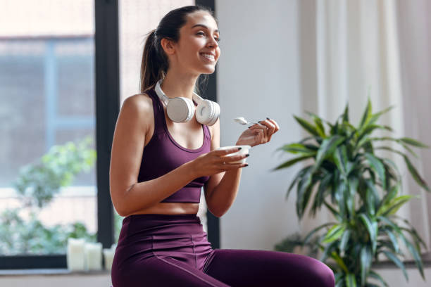 joven deportista comiendo un yogur mientras está sentada en una pelota de fitness en casa. - salud y belleza fotografías e imágenes de stock