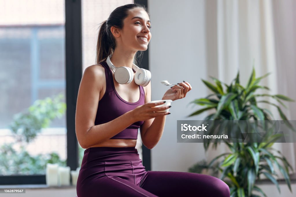 Sporty young woman eating a yogurt while sitting on fitness ball at home. Shot of sporty young woman eating a yogurt while sitting on fitness ball at home. Women Stock Photo