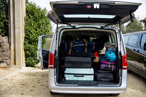 An open rental van boot filled with suitcases and luggage, loaded and ready for the return home after a holiday. The van is parked on a gravel driveway at a villa in Castelferrus, France.