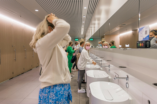 Inside the women's bathroom in an airport, a group of women standing at the sinks and mirrors, freshening themselves up before getting on a flight back home. Some are washing their hands and one woman is putting her hair up. They are all wearing protective face masks, to reduce the spread of COVID-19.
