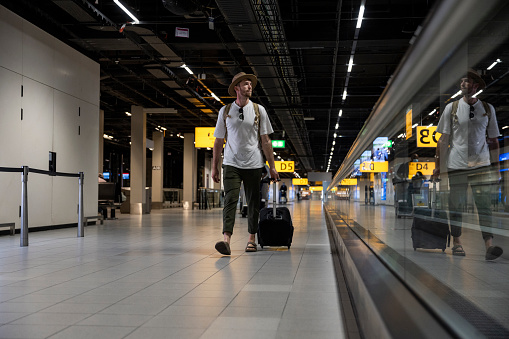 A young backpacker pulling a small suitcase behind him while walking through the airport in the Netherlands. He is looking to his side as he walks.