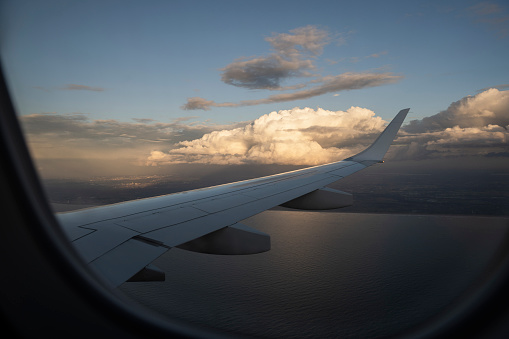 Photographed through a window, an airplane wing flying above water, next to a cloudy sky during dusk..
