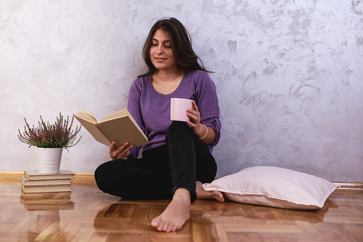 Wide shot of smiling young woman sitting on the floor, by the wall, enjoying a cup of coffee and reading an interesting book.