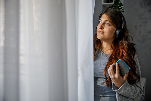 Copy space shot of smiling young woman standing by the window, looking outside, contemplating and listening to music via headphones.