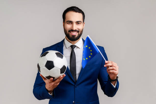 Happy man holds black and white soccer ball and flag of EU watching football support favourite team. Happy bearded man holding black and white soccer ball and flag of EU, watching football and support favourite team, wearing official style suit. Indoor studio shot isolated on gray background. celebrity endorsements stock pictures, royalty-free photos & images