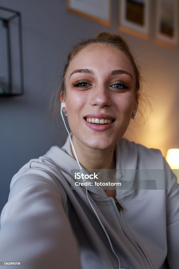Joyful teenage girl looking at camera as if she is on a video call Front view of joyful teenage girl, wearing in-ear headphones, smiling at camera as if she is holding a smart phone during a video call with a friend. Selfie Stock Photo